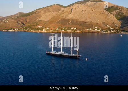 Idra, Grecia - 30 luglio 2025: Vista aerea di un maestoso yacht a vela che naviga attraverso il mare blu profondo sullo sfondo di montagne baciate dal sole e pittoresche case costiere. Foto Stock