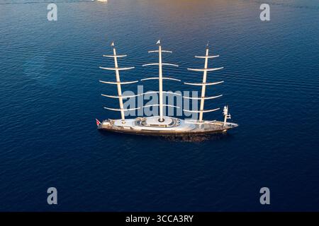 Idra, Grecia - 30 luglio 2025: Veduta aerea di un elegante yacht a vela che attraversa il mare blu profondo, i suoi alberi bianchi contrastano nettamente con lo scafo scuro e l'acqua scintillante. Foto Stock
