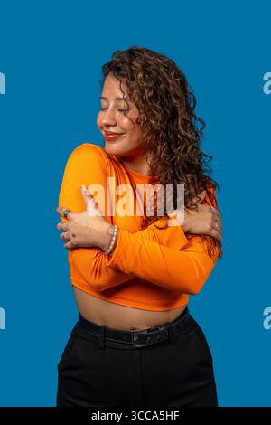 Giovane donna latina che si abbraccia con gli occhi chiusi, sente amore e auto-accettazione su sfondo blu Foto Stock