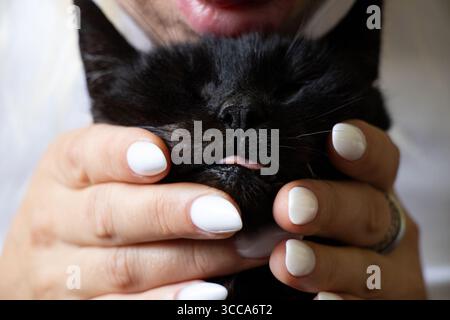 Ragazza che tiene in grembo un gatto nero, sostenendone delicatamente la museruola con una lingua allungata e un naso con un primo piano in manicure bianca Foto Stock
