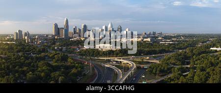 La vista aerea dello skyline di Charlotte si erge maestosamente sopra alberi verdeggianti e complessi incroci autostradali sotto un cielo sereno, Charlotte, North Caroli Foto Stock