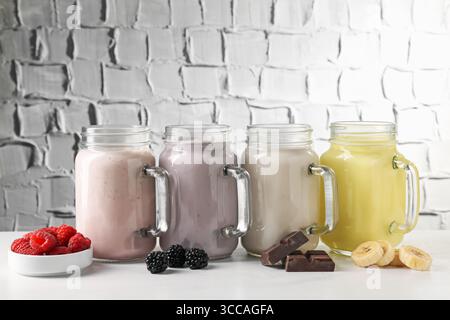 Diversi frullati gustosi nei vasetti mason e ingredienti sul tavolo bianco Foto Stock