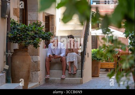 Chania, Creta, Grecia - 22 giugno 2025: Una coppia si rilassa sulle scale a Chania, Creta, godendosi il sole e i loro telefoni. Foto Stock