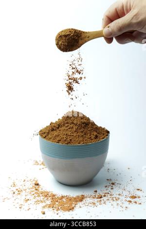 Polvere di cacao che cade dal cucchiaio di legno nel recipiente su sfondo bianco, creando una nuvola di polvere Foto Stock