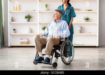 Ritratto a lunghezza intera del paziente anziano di sesso maschile in sedia a rotelle e. il suo giovane caregiver a casa di riposo Foto Stock