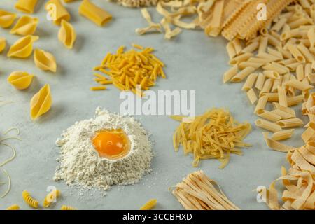 Diversi tipi di pasta che formano un cerchio intorno alla farina con un tuorlo d'uovo su sfondo grigio, pronto per essere cotto Foto Stock