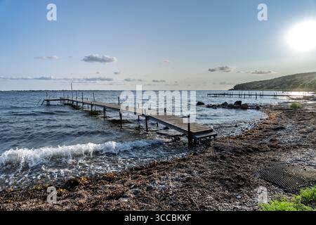 Un tranquillo scenario costiero caratterizzato da un molo di legno che si estende in dolci onde sotto un cielo soleggiato, con coste frastagliate e colline lontane nel backgroun Foto Stock