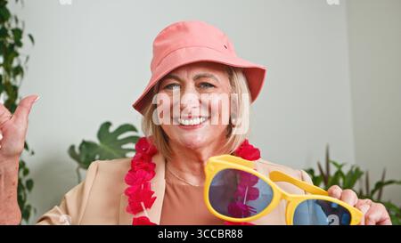 Donna anziana sorridente mentre tiene in mano occhiali da sole gialli in abito festivo Foto Stock