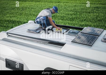 Un uomo ispeziona e pulisce i pannelli solari sul tetto di un veicolo ricreativo in un campo aperto sotto cieli limpidi, garantendo un'efficiente acquisizione di energia. Foto Stock