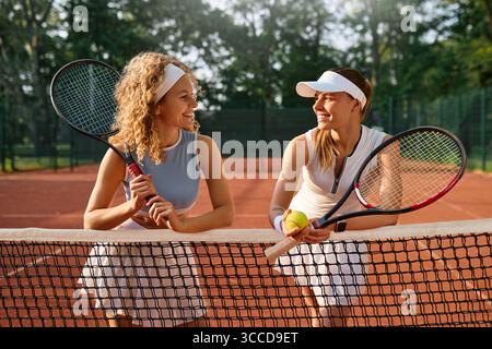 Due tenniste sorridenti in piedi a rete dopo la partita sul campo in terra battuta. Momenti di riposo e conversazione amichevole alla luce dell'estate. Concep Foto Stock