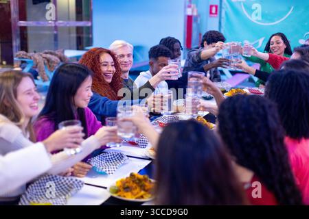 Amici multietnici felici che alzano i bicchieri d'acqua e brindano seduti a un tavolo del ristorante, festeggiando insieme Foto Stock