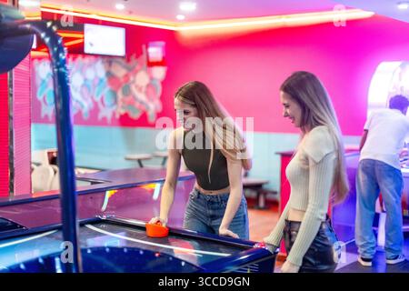 Due amiche che si divertono con una partita di hockey da tavolo, mostrando il divertimento e l'emozione dell'intrattenimento arcade Foto Stock