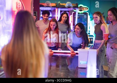 Gruppo di amici che si divertono a giocare a hockey da tavolo in una vivace sala giochi illuminata, piena di risate ed emozioni Foto Stock