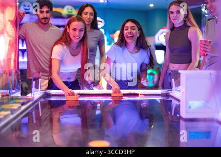 Un gruppo di giovani amici che giocano a hockey da tavolo in una sala giochi colorata e si divertono insieme Foto Stock