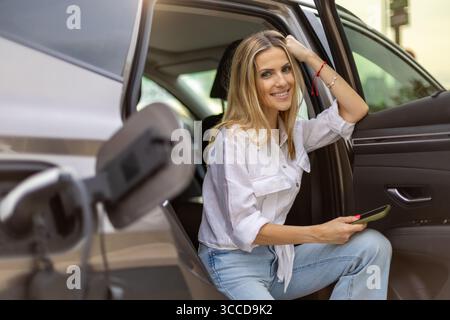 Donna d'affari sorridente che esce dalla sua auto elettrica tenendo in mano uno smartphone, incarna la tecnologia moderna e il trasporto sostenibile Foto Stock