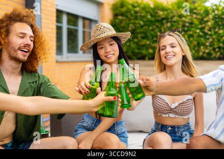 Un gruppo di allegri amici multietnici che brindano con bottiglie di birra mentre si godono le feste estive in un cortile della casa Foto Stock