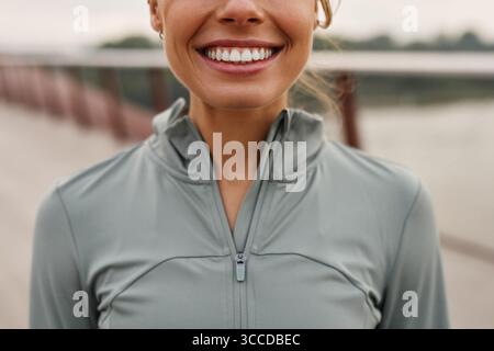 Una giovane donna allegra con una giacca atletica verde chiaro sorride con fiducia su un ponte panoramico, mostrando il suo impegno per una vita attiva e sana Foto Stock