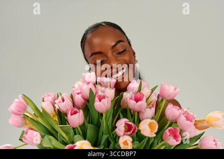 Donna afroamericana con bouquet di tulipani rosa, che celebra la festa della mamma o della donna con gioia e amore, sorridendo brillantemente su sfondo neutro. Conc Foto Stock