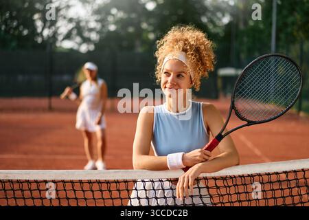La giovane donna sorridente in abbigliamento sportivo si appoggia alla rete da tennis, tenendo la racchetta in sicurezza dopo una partita su un campo di terra battuta. Concetto di fiducia, empowerment, a Foto Stock