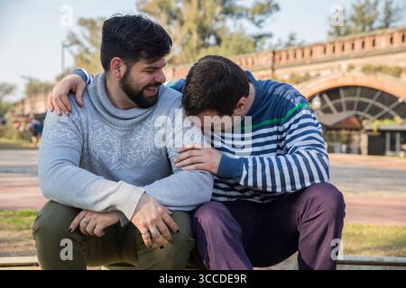 Due uomini si siedono vicini in un parco, condividendo risate e sorrisi caldi in un pomeriggio luminoso. Foto Stock