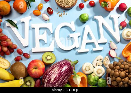 La parola VEGAN in grandi lettere bianche su una superficie blu, circondata da frutta fresca, verdure e una ciotola di lenticchie, dieta vegana piatta Foto Stock