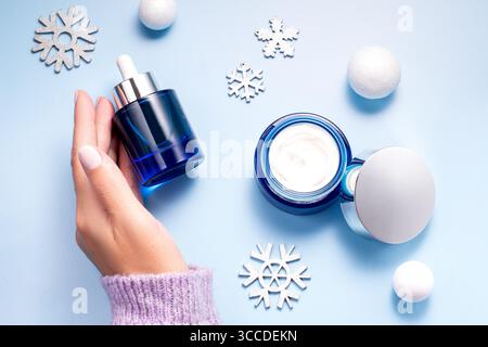 Concetto di bellezza invernale per la cura della pelle con mani di donna che tengono il siero per il viso e crema idratante su sfondo blu, vista dall'alto Foto Stock