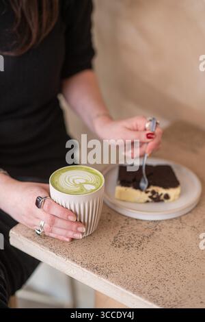 Primo piano di una mano che tiene una tazza di latte matcha in ceramica mentre usa una forchetta per prendere un boccone di torta al cioccolato in un accogliente bar. Foto Stock