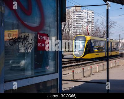 Kiev, Ucraina -6 marzo 2025: Moderno tram giallo che si avvicina alla stazione in Ucraina, passando vicino ad una vecchia finestra sporca con graffiti e un cartello affittato, Foto Stock