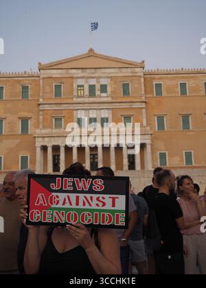 Atene, Grecia - 10 agosto 2025: Persona in possesso di un cartello che protesta contro il genocidio di fronte a grandi edifici, probabile struttura governativa Foto Stock