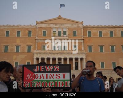 Atene, Grecia - 10 agosto 2025: I manifestanti tengono il cartello "ebrei contro il genocidio" di fronte all'edificio del parlamento greco con la bandiera Foto Stock
