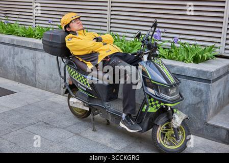 Addetto alla consegna dei pacchi che riposa su uno scooter su un marciapiede a Pechino, in Cina, il 21 aprile 2024 Foto Stock