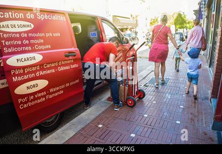 Le Touquet, Francia - 22 agosto 2023: Un furgone rosso che espone specialità locali è parcheggiato su un marciapiede di mattoni mentre un uomo scarica merci mentre le famiglie con Foto Stock