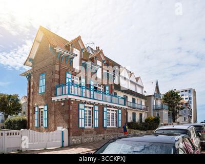 Le Touquet, Francia - 22 agosto 2023: Una pittoresca casa in mattoni con persiane blu vibranti e un cancello bianco si erge sotto un cielo parzialmente nuvoloso sulla costa Foto Stock
