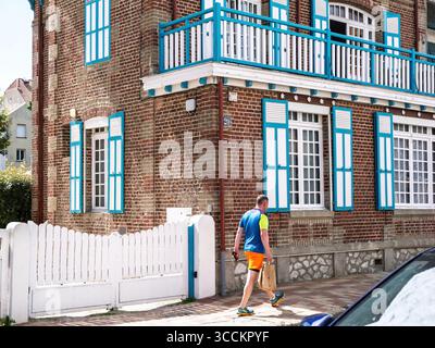 Le Touquet, Francia - 22 agosto 2023: Un uomo in un abbigliamento sportivo luminoso passa davanti a un'affascinante casa in mattoni con persiane blu vibranti e un cancello per picchetti bianco Foto Stock