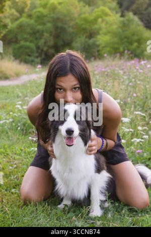 Giovane donna indiana inginocchiata dietro il cane Border Collie nel prato, entrambe guardano la macchina fotografica con un sorriso. Ideale per i temi della cura degli animali domestici e del legame uomo-animale Foto Stock
