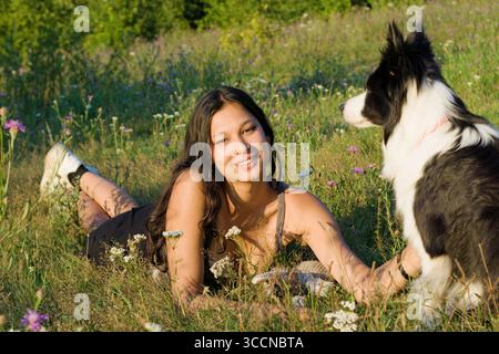 Giovane donna indiana sdraiata sul suo stomaco in erba giocando con un cane Border Collie in una giornata estiva di sole. stile di vita, cura degli animali domestici, pet sitting Foto Stock