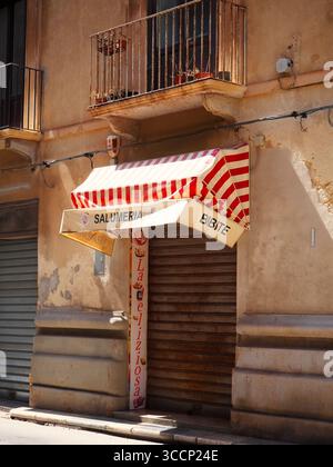 Arrugginito di un piccolo negozio di alimentari con tenda a righe rosse in una stradina stretta del centro storico di Trapani, in Sicilia, alla luce del sole estivo Foto Stock