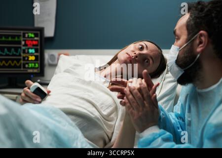 Caucasica giovane donna adulta sdraiata nel letto d'ospedale tenendo le mani con Caucasico giovane adulto uomo che indossa maschera medica e abito, monitor medico che visualizza i segni vitali in background Foto Stock