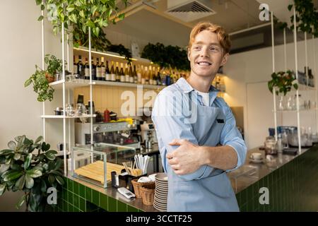 Allegro barista in piedi tranquillamente con le braccia piegate e il grembiule in un caldo ambiente da caffè Foto Stock