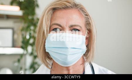 Medico donna anziano che indossa una maschera medica e sorride in Bright Clinic Foto Stock