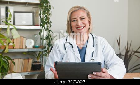 Sorridente Senior Doctor Holding tablet in accogliente ambiente di ufficio medico Foto Stock