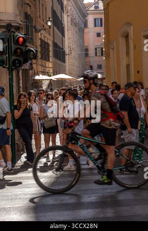 Un ciclista aspetta ad un incrocio su una trafficata strada di Roma, circondato da pedoni pronti ad attraversare, con traffico e vita cittadina animata sullo sfondo. Foto Stock