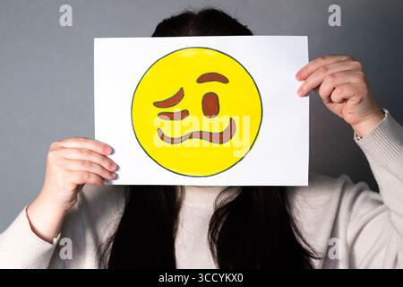 Donna che tiene confusa maschera emoji davanti al viso. Tendenze dei social media, marketing Gen Z, giornata mondiale delle emoji 17 luglio. Foto Stock
