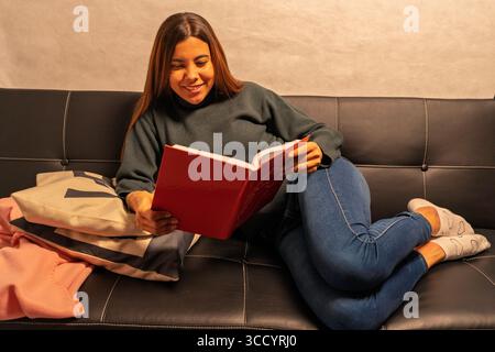 Una giovane donna gode di un momento tranquillo di svago, leggendo comodamente un libro sul suo divano Foto Stock
