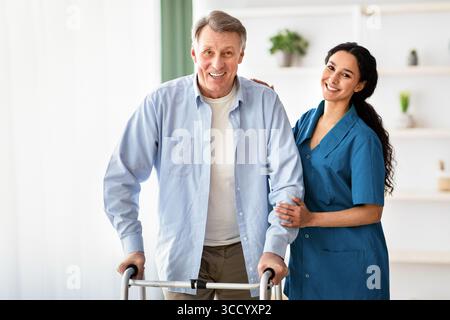 Ritratto di un uomo anziano felice che usa un camminatore e una bella giovane infermiera che lo aiuta a casa di riposo. Servizio di assistenza per anziani Foto Stock
