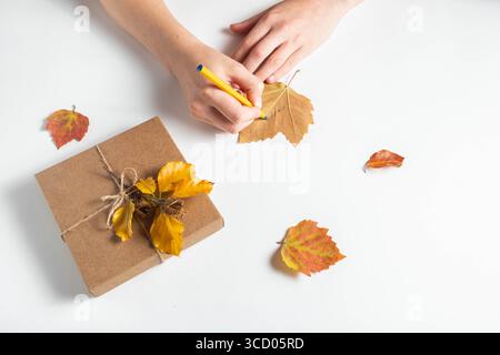Le mani femminili scrivono su una foglia d'acero per decorare una confezione regalo con foglie autunnali su sfondo bianco. Concetto di Ringraziamento e zero rifiuti Foto Stock