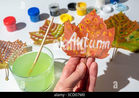 La mano della donna tiene un foglio di autunno con l'iscrizione Autumn. Pitture di gouache e un pennello sul tavolo. Processo creativo e concetto di zero sprechi Foto Stock