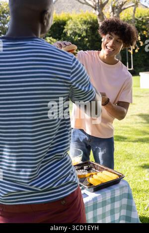 Diversi amici maschi scambiano hamburger su un tavolo da picnic con vassoio di mais e ciotola nel cortile posteriore Foto Stock