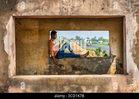 Bogura, Bangladesh - 4 giugno 2024: Veduta di un giovane uomo si siede contemplativamente in una cornice di finestra intemprata, guardando fuori un'autostrada, texture contrastanti di vecchia pietra e vibrante vegetazione. Foto Stock