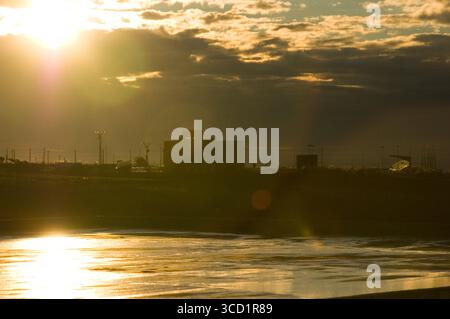 Un paesaggio sagomato è immerso nel caldo bagliore del sole che tramonta, con un corpo d'acqua in primo piano che riflette la luce. Foto Stock
